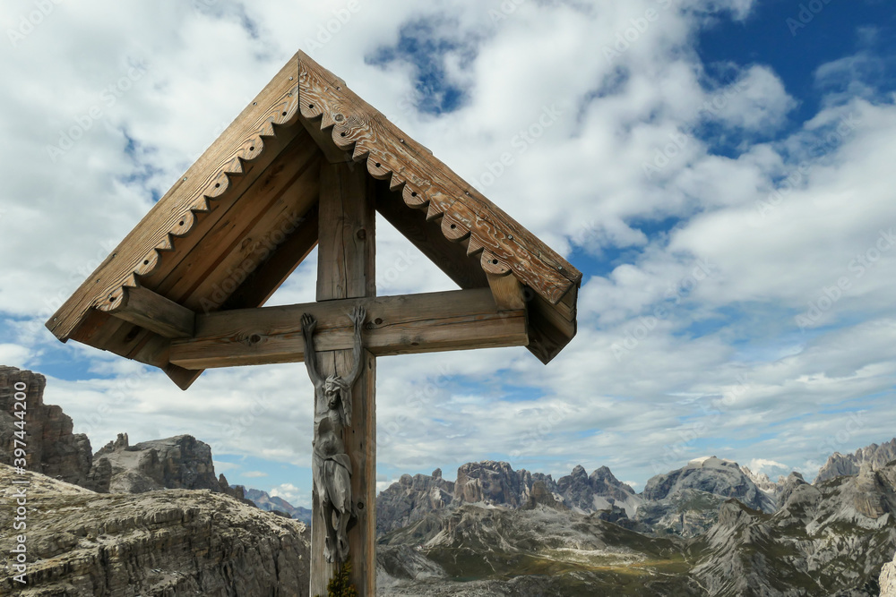 A wooden cross with a figure of Jesus on it with panoramic view on Dolomites in Italy. There are sharp and steep mountain slopes around. The sky is full of soft clouds. Spirituality and meditation A wooden cross with a figure of Jesus on it with panoramic view on Dolomites in Italy. There are sharp and steep mountain slopes around. The sky is full of soft clouds. Spirituality and meditation