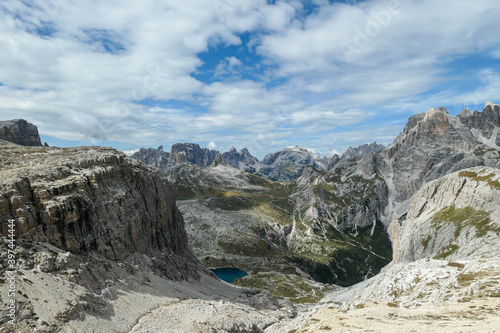 A panoramic view on Dolomites in Italy. There are sharp and steep mountain slopes around. At the bottom of a small valley there is a small navy blue lake. The sky is full of soft clouds. Raw landscape