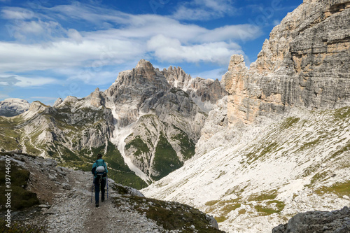 A woman with backpack and sticks hiking on a path in Italian Dolomites. There are sharp and steep mountains around. Lower slopes of the mountains overgrown with small, green plants. Raw landscape