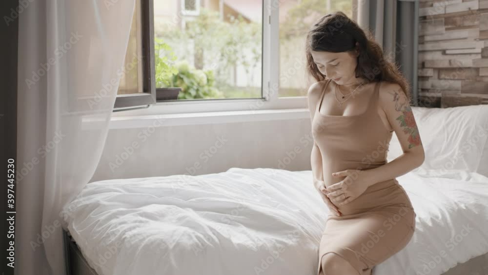 Happy pregnant woman stroking belly near window indoors. Pretty expectant mother sitting in light room. Future mom petting her stomach. Pregnant woman smiling to unborn baby Happy pregnant woman stroking belly near window indoors. Pretty expectant mother sitting in light room. Future mom petting her stomach. Pregnant woman smiling to unborn baby