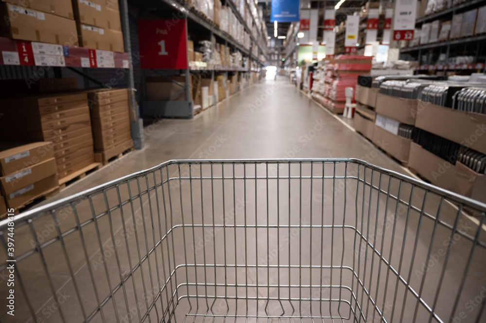 Empty shopping cart in a furniture store warehouse. Supermarket shopping basket aisle with box shelf abstract blur defocused background Empty shopping cart in a furniture store warehouse. Supermarket shopping basket aisle with box shelf abstract blur defocused background