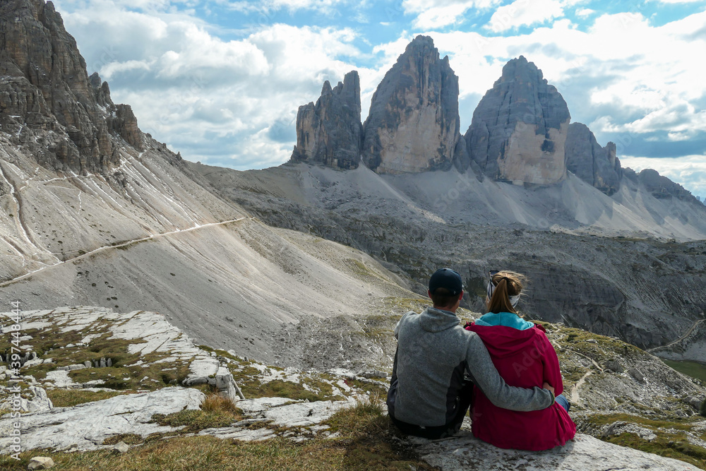A couple in hiking outfit, sitting on the ground and enjoying the view on the famous Tre Cime di Lavaredo (Drei Zinnen) in Italian Dolomites. Desolated and raw landscape, full of lose stones. Overcast