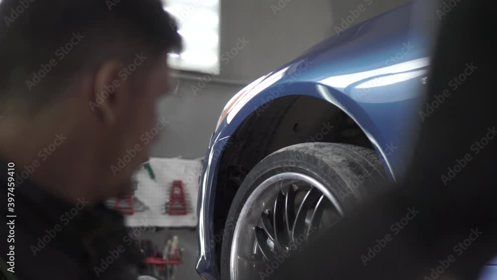 Closeup view of mechanic lifts the car in the service using a lift. Footage. Over the shoulder, blurred silhouette of a repairman preparing to carry out repair operations in a workshop. Closeup view of mechanic lifts the car in the service using a lift. Footage. Over the shoulder, blurred silhouette of a repairman preparing to carry out repair operations in a workshop.