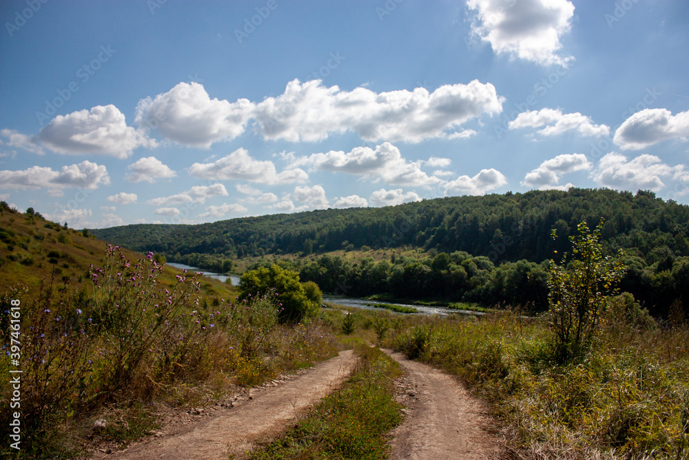 Dirt winding road to the river on a sunny day. Summer landscape on a clear sunny day. Wildlife in the summer in central Russia. White clouds in the blue sky over a dirt road.