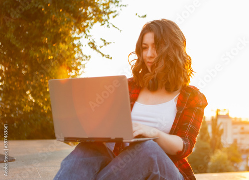 Young cheerful woman student sitting with laptop outdoors early in the morning at sunrise
