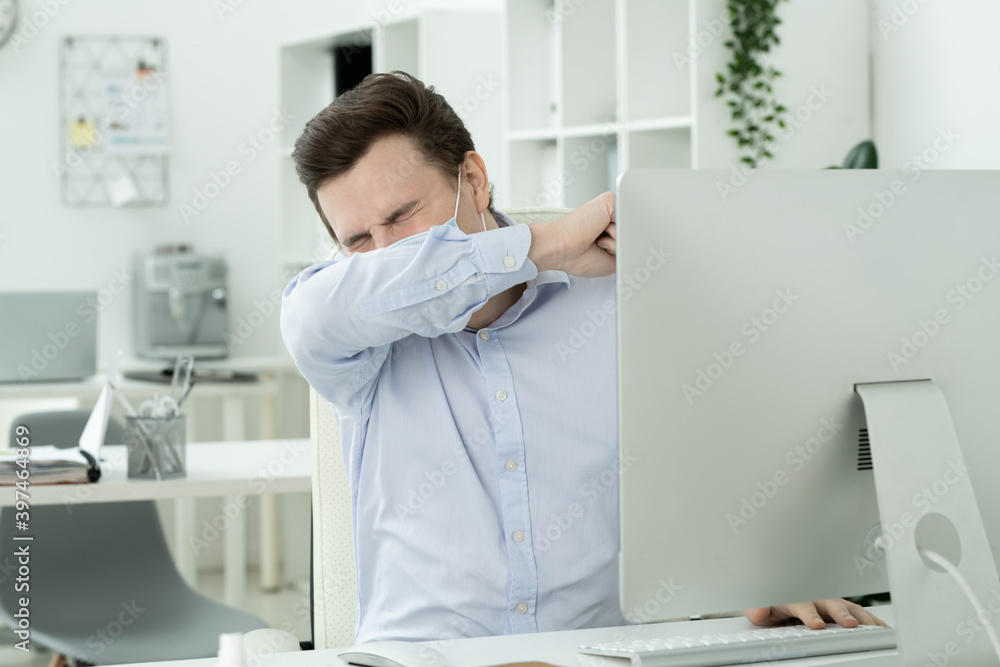 Young manager in protective mask putting face on arm while sneezing by workplace