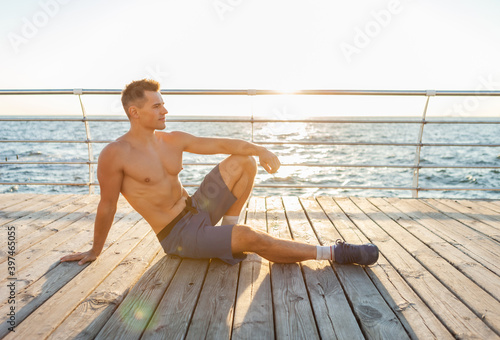 Tired muscular handsome fitness man with naked torso resting while sitting on the beach at sunrise © splitov27