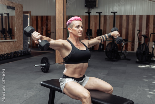 Young athletic strong woman trains the muscles of the shoulder with dumbbells in her hands while sitting on a bench in a cross gym. Bodybuilding and Fitness