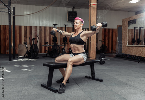 Young athletic strong woman trains the muscles of the shoulder with dumbbells in her hands while sitting on a bench in a cross gym. Bodybuilding and Fitness