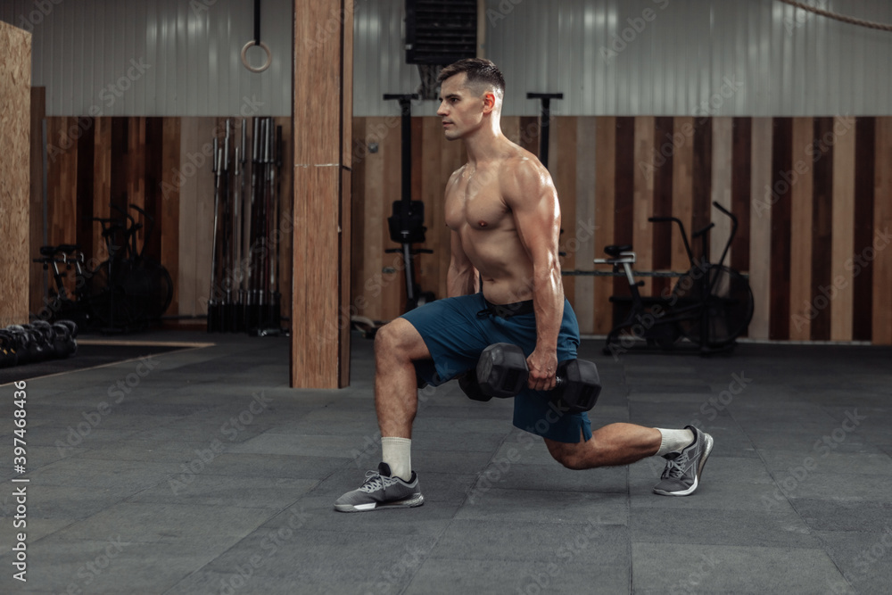 Muscular man training his legs, doing lunges with heavy dumbbells in modern gym. Healthy lifestyle concept