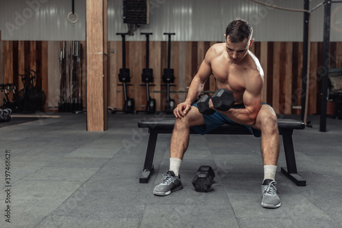 Athletic man exercising with heavy dumbbell  concentrated training biceps while sitting on a bench in the gym. Bodybuilding and Fitness