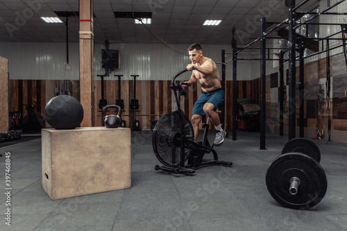 Powerful muscular male athlete with a naked torso trains with an air bike in a modern health club. Functional, cross fit training. Cardio exercise. Healthy lifestyle concept