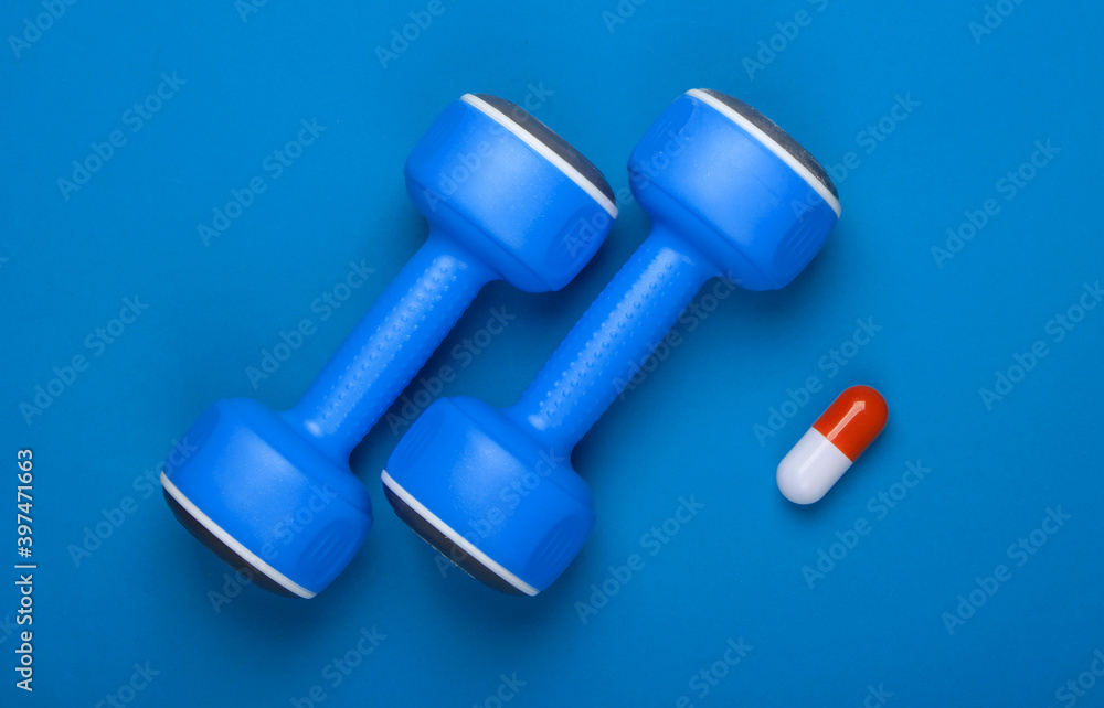Pharmacology in sports. Dumbbell and capsule on blue background. Vitamins, steroids. Top view Pharmacology in sports. Dumbbell and capsule on blue background. Vitamins, steroids. Top view
