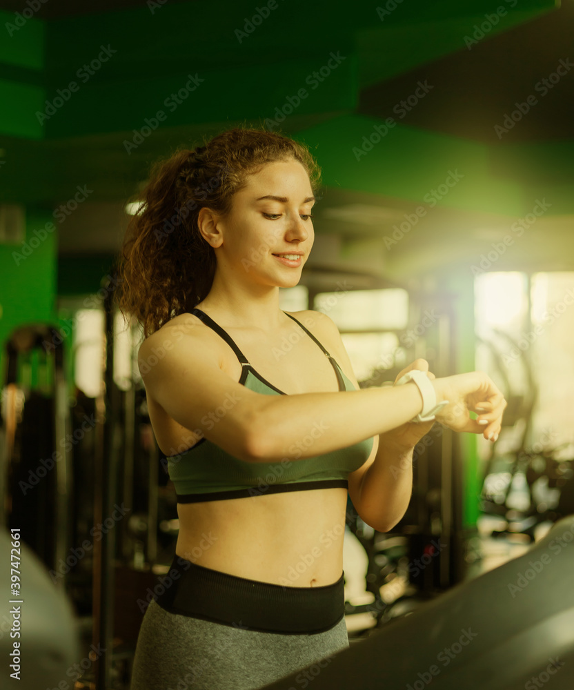 Fototapeta premium Fit woman in sportswear runs on a treadmill and looks at her watch in the gym. The concept of a healthy lifestyle, warming up, fitness, weight loss.