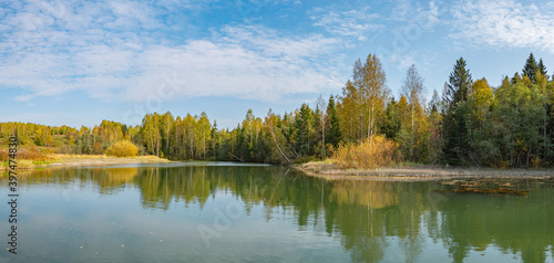 Autumn forest trees are reflected in the river water of the panoramic landscape. Blue sky with clouds.