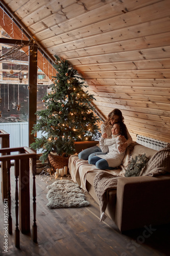 Young mother with two little daughters in pajamas decorate a New Year's tree in the cozy room with with big light window