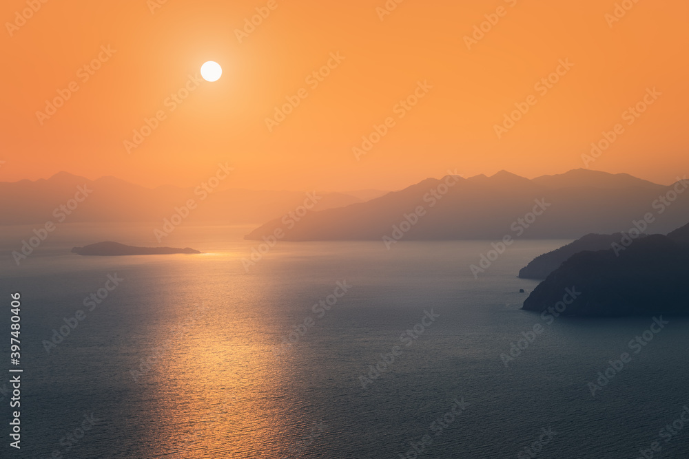 Fototapeta premium Moody sunset, the distant sun sets behind the silhouettes of blue mountains on the horizon, washed by the waters of the Mediterranean sea