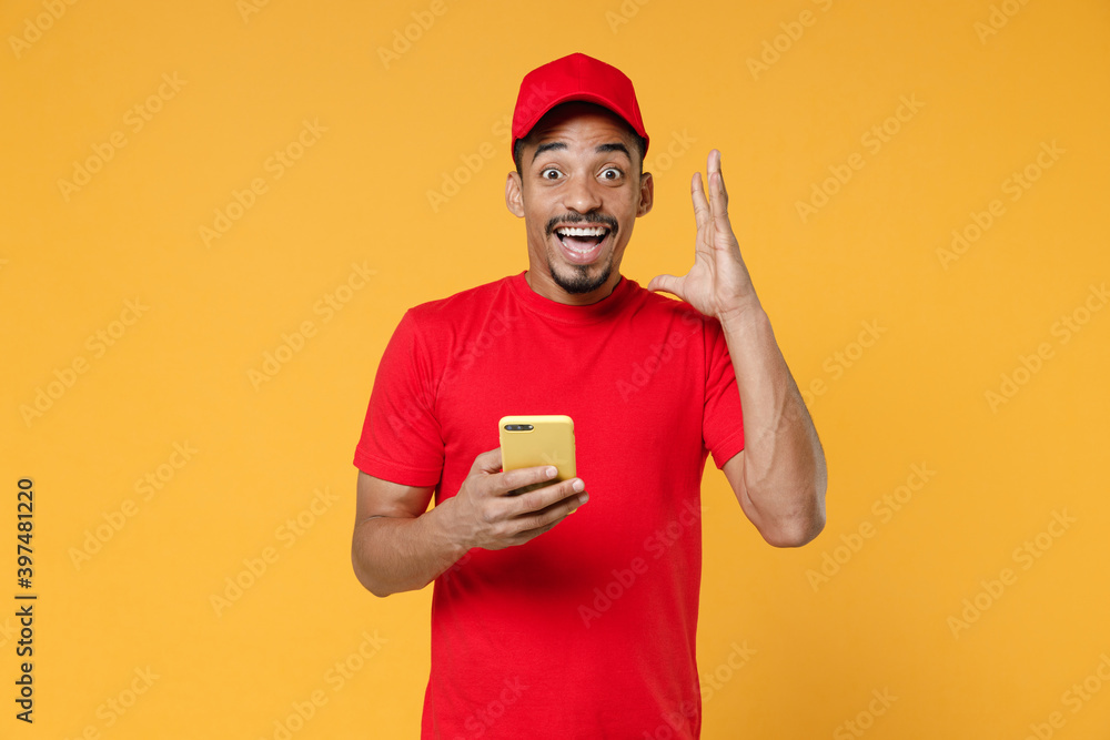 Smiling delivery employee african man 20s in red cap blank print t-shirt uniform workwear work courier dealer service concept hold in hand using mobile cell phone isolated on yellow background studio. Smiling delivery employee african man 20s in red cap blank print t-shirt uniform workwear work courier dealer service concept hold in hand using mobile cell phone isolated on yellow background studio.