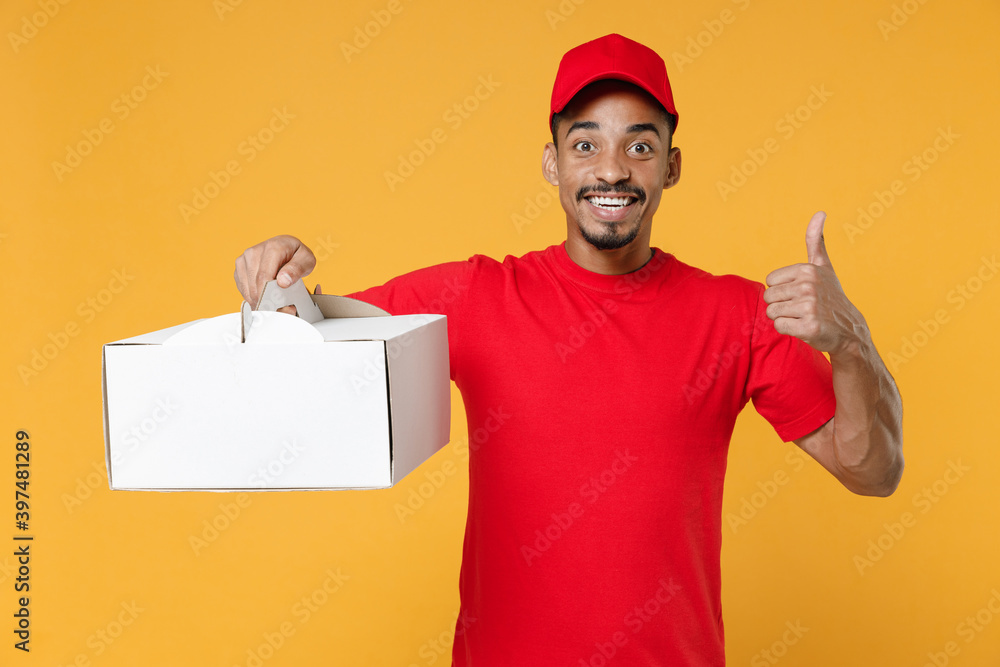 Delivery employee african man 20s in red cap blank print t-shirt uniform workwear work courier dealer service concept hold cake dessert in unmarked cardboard box isolated on yellow background studio.