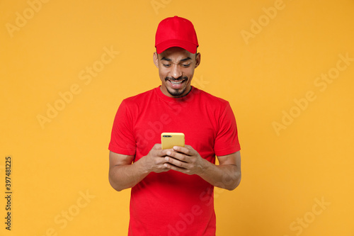 Smiling delivery employee african man 20s in red cap blank print t-shirt uniform workwear work courier dealer service concept hold in hand using mobile cell phone isolated on yellow background studio.