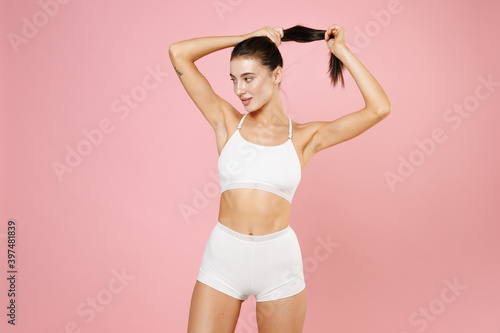 Smiling beautiful pretty young brunette woman 20s in white underwear with sexy body standing posing put hands on head making ponytail isolated on pastel pink colour background, studio portrait.