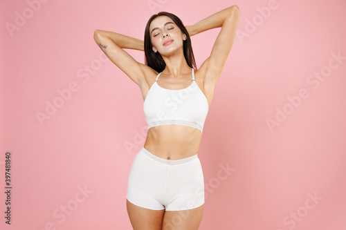 Seduction tender beautiful young brunette woman in white underwear standing with hands up showing result on underarms removing hair procedure isolated on pastel pink colour background studio portrait. © ViDi Studio