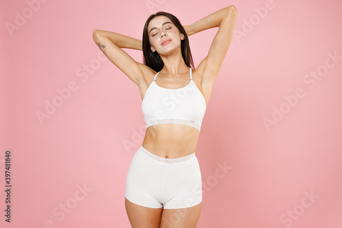 Seduction tender beautiful young brunette woman in white underwear standing with hands up showing result on underarms removing hair procedure isolated on pastel pink colour background studio portrait.