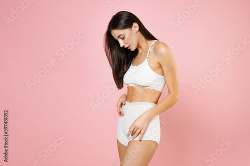 Side view of attractive seductive young brunette woman 20s in white underwear with beautiful body posing looking hold hands on stomach belly isolated on pastel pink colour background, studio portrait. © ViDi Studio