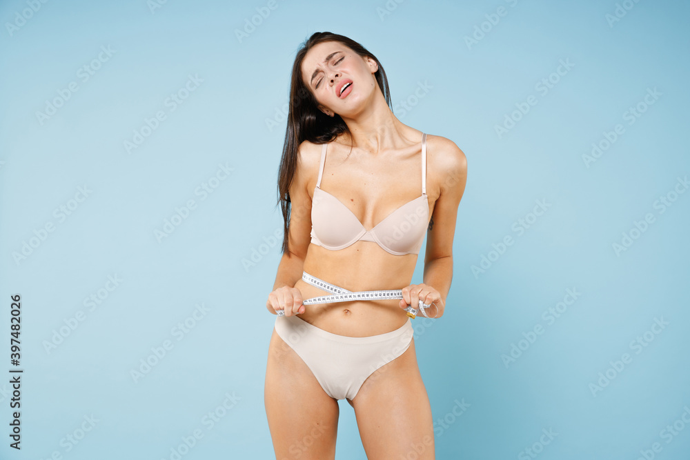 Upset displeased dissatisfied young brunette woman 20s in beige brassiere underwear standing posing measuring waist with measure tape isolated on pastel blue colour wall background studio portrait. Upset displeased dissatisfied young brunette woman 20s in beige brassiere underwear standing posing measuring waist with measure tape isolated on pastel blue colour wall background studio portrait.