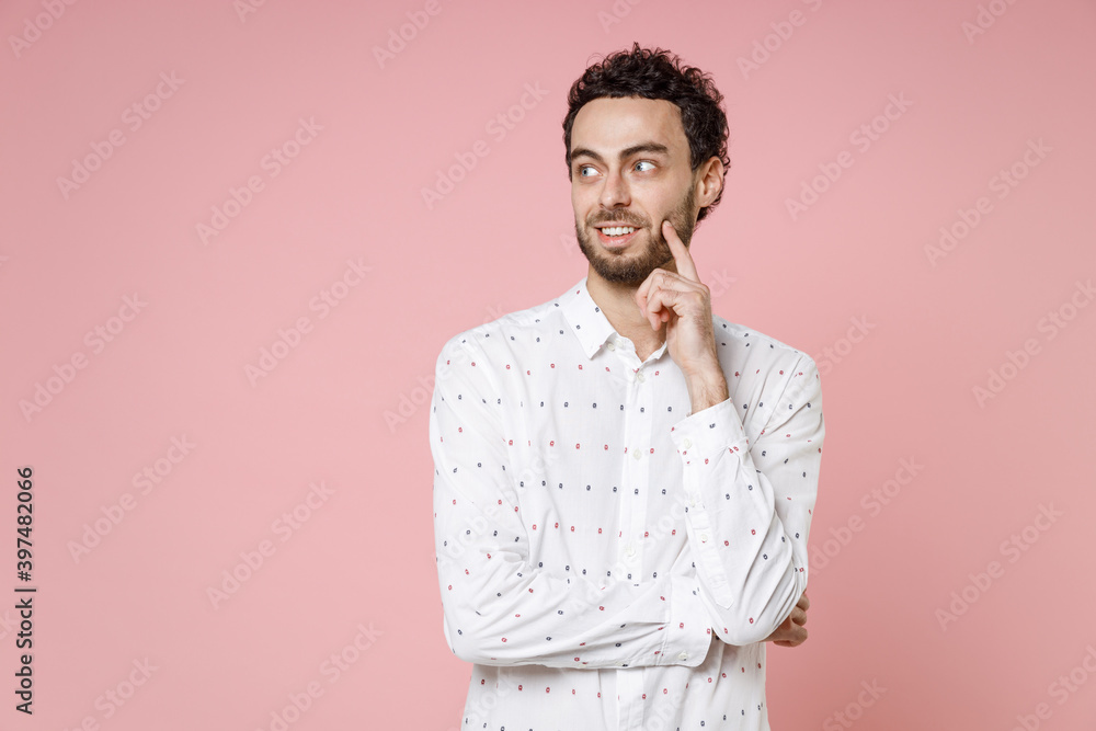 Smiling pensive handsome young bearded man 20s wearing basic casual white shirt standing put hand prop up on chin looking aside isolated on pastel pink color wall background studio portrait. Smiling pensive handsome young bearded man 20s wearing basic casual white shirt standing put hand prop up on chin looking aside isolated on pastel pink color wall background studio portrait.