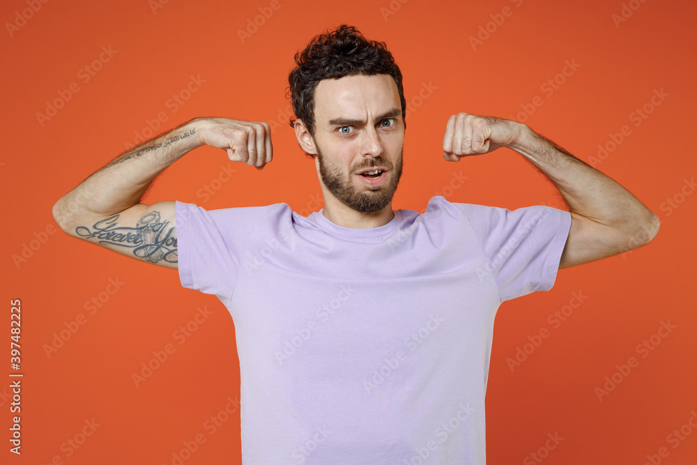Strong shocked cheerful young bearded man 20s wearing basic casual violet t-shirt standing showing biceps muscles looking camera isolated on bright orange color wall background studio portrait. Strong shocked cheerful young bearded man 20s wearing basic casual violet t-shirt standing showing biceps muscles looking camera isolated on bright orange color wall background studio portrait.