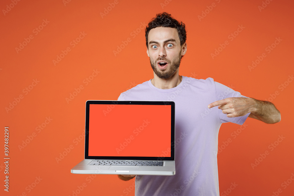 Shocked young bearded man 20s wearing casual basic violet t-shirt standing pointing index finger on laptop pc computer with blank empty screen isolated on orange color wall background studio portrait. Shocked young bearded man 20s wearing casual basic violet t-shirt standing pointing index finger on laptop pc computer with blank empty screen isolated on orange color wall background studio portrait.