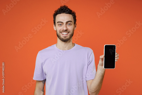 Cheerful smiling funny young bearded man 20s in casual violet t-shirt standing hold mobile cell phone with blank empty screen looking camera isolated on bright orange color background studio portrait.
