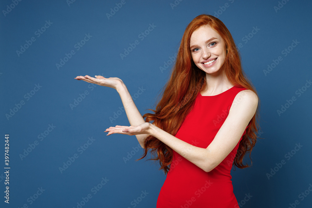 Smiling cheerful beautiful young redhead woman 20s wearing bright red elegant evening dress standing pointing hands aside on mock up copy space isolated on blue color wall background studio portrait. Smiling cheerful beautiful young redhead woman 20s wearing bright red elegant evening dress standing pointing hands aside on mock up copy space isolated on blue color wall background studio portrait.