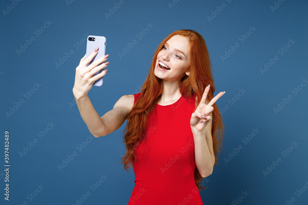 Cheerful funny young redhead woman 20s wearing bright red elegant evening dress standing doing selfie shot on mobile phone showing victory sign isolated on blue color wall background studio portrait. Cheerful funny young redhead woman 20s wearing bright red elegant evening dress standing doing selfie shot on mobile phone showing victory sign isolated on blue color wall background studio portrait.