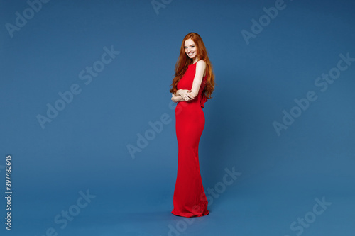 Full length side view of smiling young redhead woman 20s wearing bright red elegant evening dress standing holding hands crossed looking camera isolated on blue color wall background studio portrait.