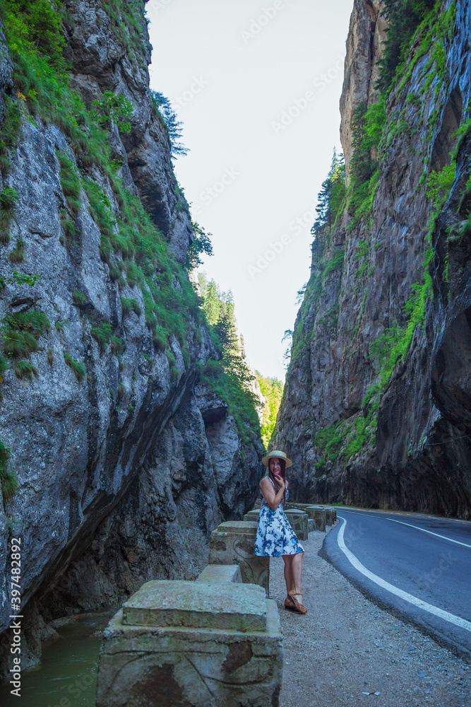 Elegant woman dressed in white blue dress and straw hat sitting near mountain river and dangerous road in Bicaz Canyon. Bicaz Key, geological wonders of nature, Transylvania, Romania.