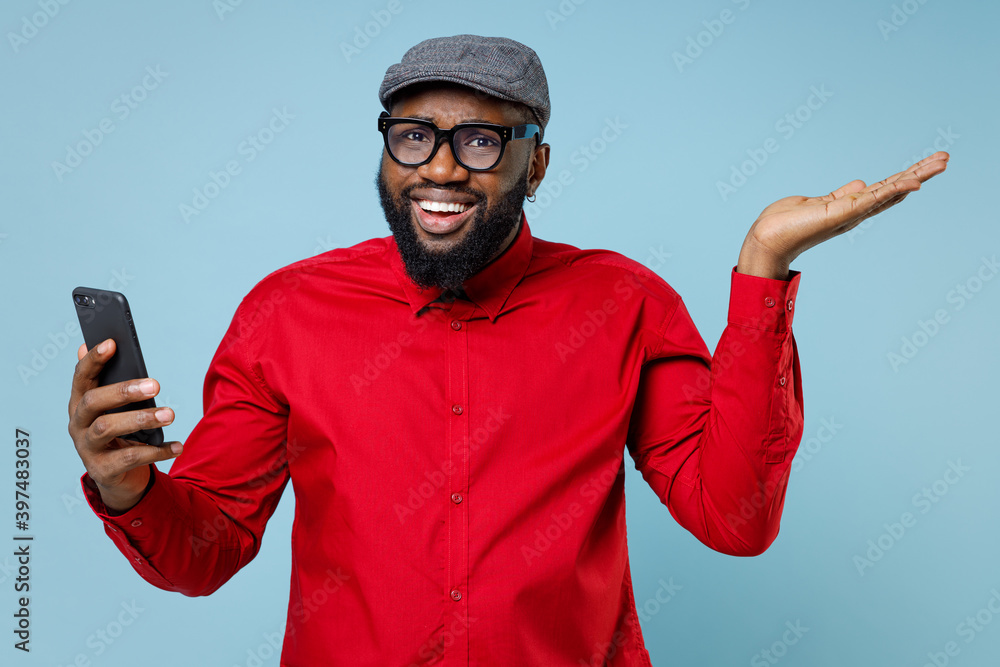 Fototapeta premium Amazed young bearded african american man 20s in casual red shirt eyeglasses cap spreading hands using mobile cell phone typing sms message isolated on pastel blue color background studio portrait.