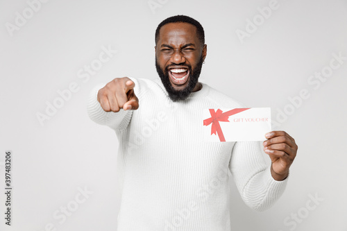 Cheerful laughing young african american man 20s wearing casual basic sweater standing hold gift certificate pointing index finger on camera isolated on white color wall background studio portrait. © ViDi Studio