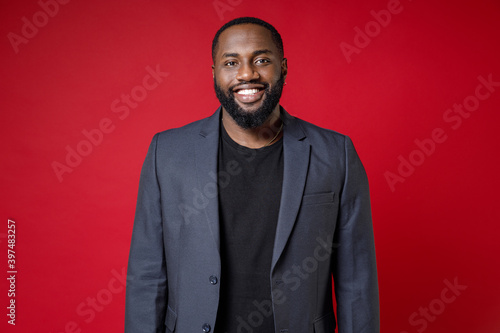 Smiling attractive confident successful young african american business man 20s wearing classic jacket suit standing and looking camera isolated on bright red color wall background studio portrait.