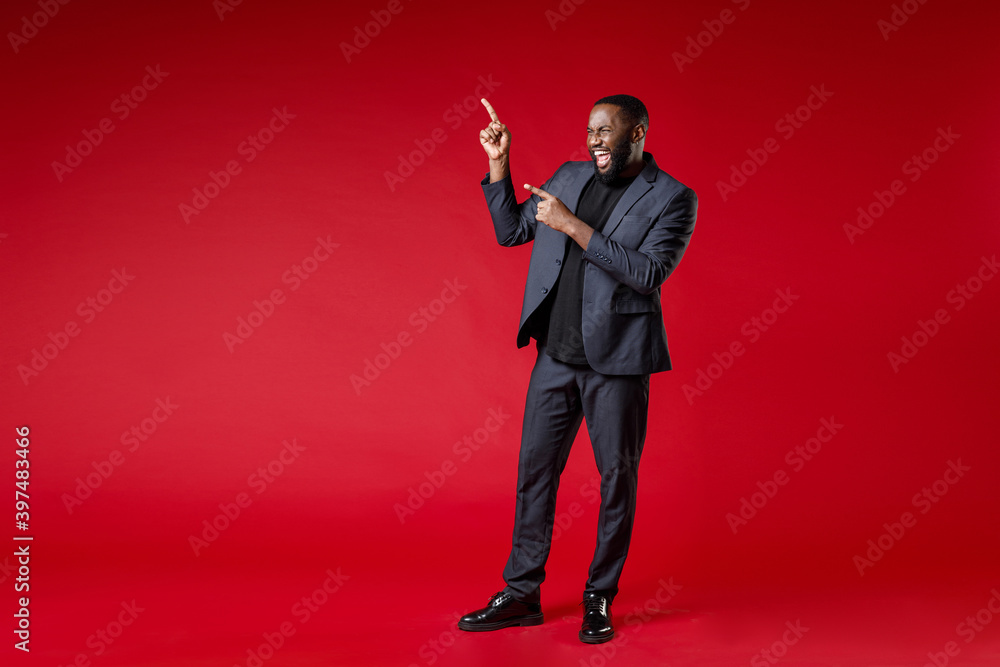 Full length of cheerful laughing young african american business man 20s wearing classic jacket suit standing pointing index fingers aside isolated on bright red color wall background studio portrait. Full length of cheerful laughing young african american business man 20s wearing classic jacket suit standing pointing index fingers aside isolated on bright red color wall background studio portrait.