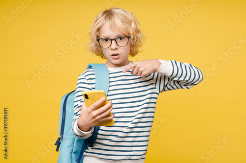 Shocked male teen boy 10s in striped sweatshirt eyeglasses backpack point index finger on mobile cell phone typing sms message isolated on yellow background, child studio portrait. Education concept.