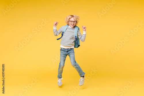 Full length of happy male boy 10s years old wearing striped sweatshirt eyeglasses backpack jumping doing winner gesture isolated on yellow color background child studio portrait. Education concept.