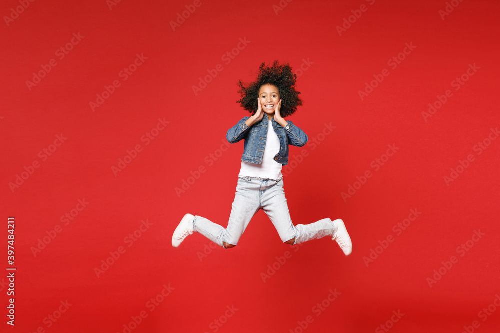 Full length of smiling little african american kid girl 12-13 years old in denim jacket jumping put hands on cheeks isolated on red background children studio portrait. Childhood lifestyle concept.
