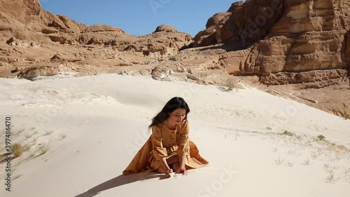 Pretty woman grabs a handful of dry sand and lets it fall between her fingers. Young traveler sitting in sunny desert dune and sifting particles fronm her hands. photo