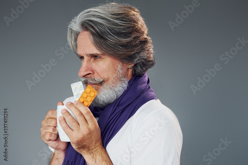 Getting sick. Against grey background. Stylish modern senior man with gray hair and beard is indoors