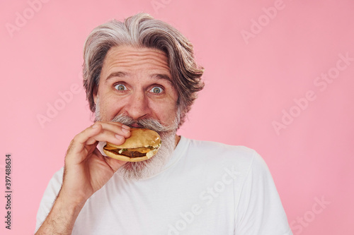 Eats delicious burger. Stylish modern senior man with gray hair and beard is indoors
