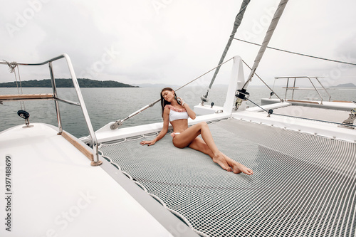 Adorable young fit lady in a white bikini with flowers is sitting on a yacht polyester net and touching her neck