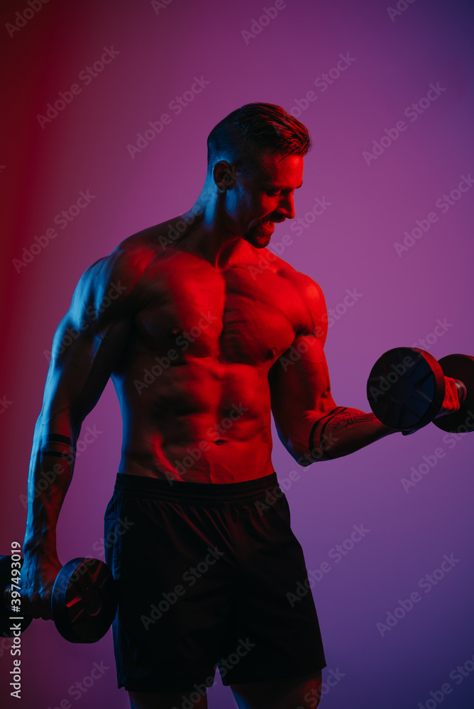 A muscular man with a beard who is doing bicep curls with dumbbells under blue and red lights. An athletic guy is demonstrating his sporty torso. An athlete is training his arms hard