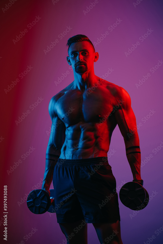 A muscular man with a beard who is posing with dumbbells under blue and red lights. An athletic guy is demonstrating his sporty torso. An athlete in shorts after biceps workout.